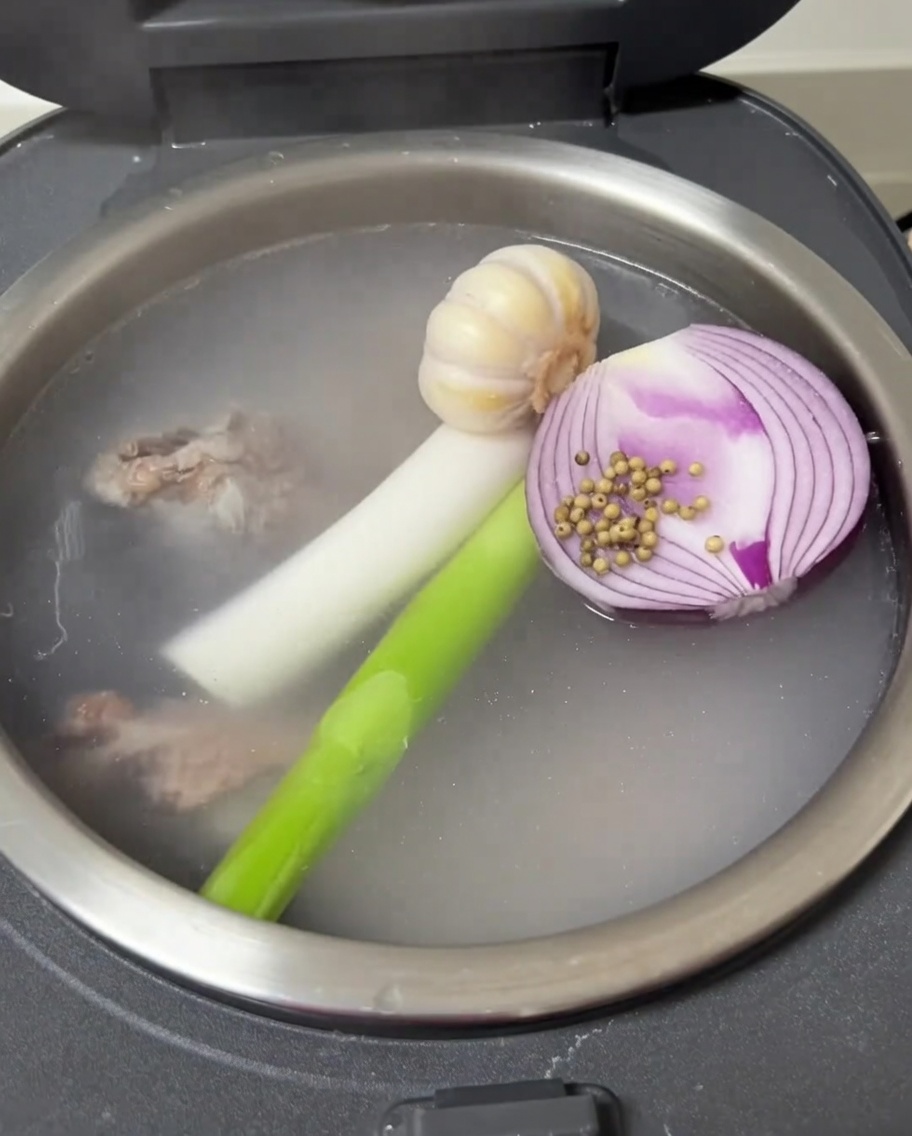 Vista del interior de la olla arrocera con ajo, cebolla, cebolleta y pimienta blanca flotando junto a las costillas.