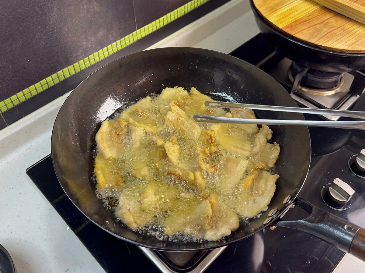 Varias láminas de panceta friéndose en un wok, siendo volteadas con pinzas de metal.