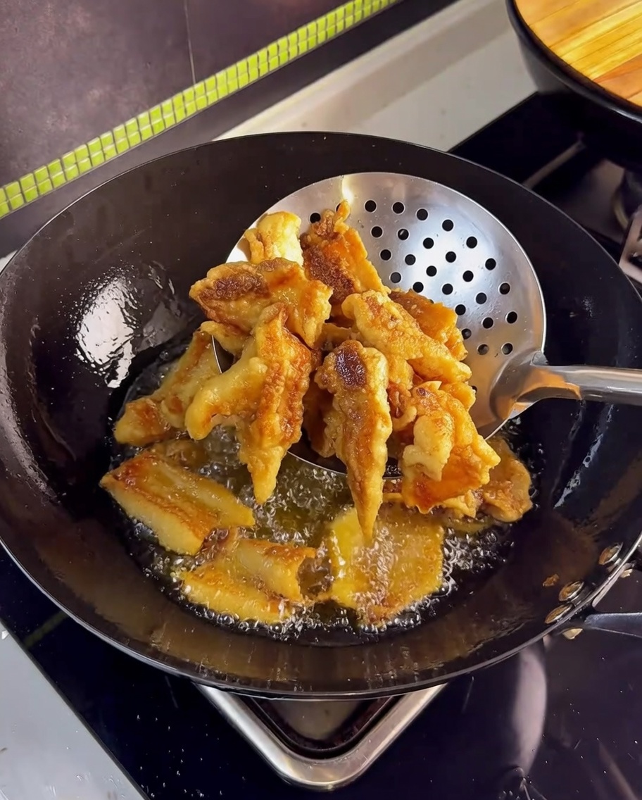 Panceta dorada y crujiente siendo retirada del aceite caliente con una espumadera de malla de acero inoxidable.