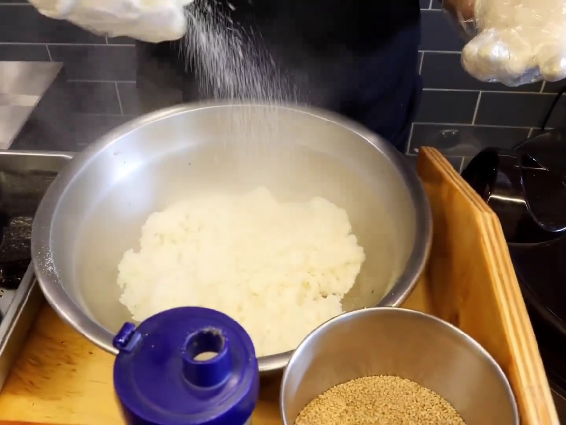 Primer plano de un bol grande de acero inoxidable lleno de arroz pegajoso humeante, con sal y semillas de sésamo por encima.