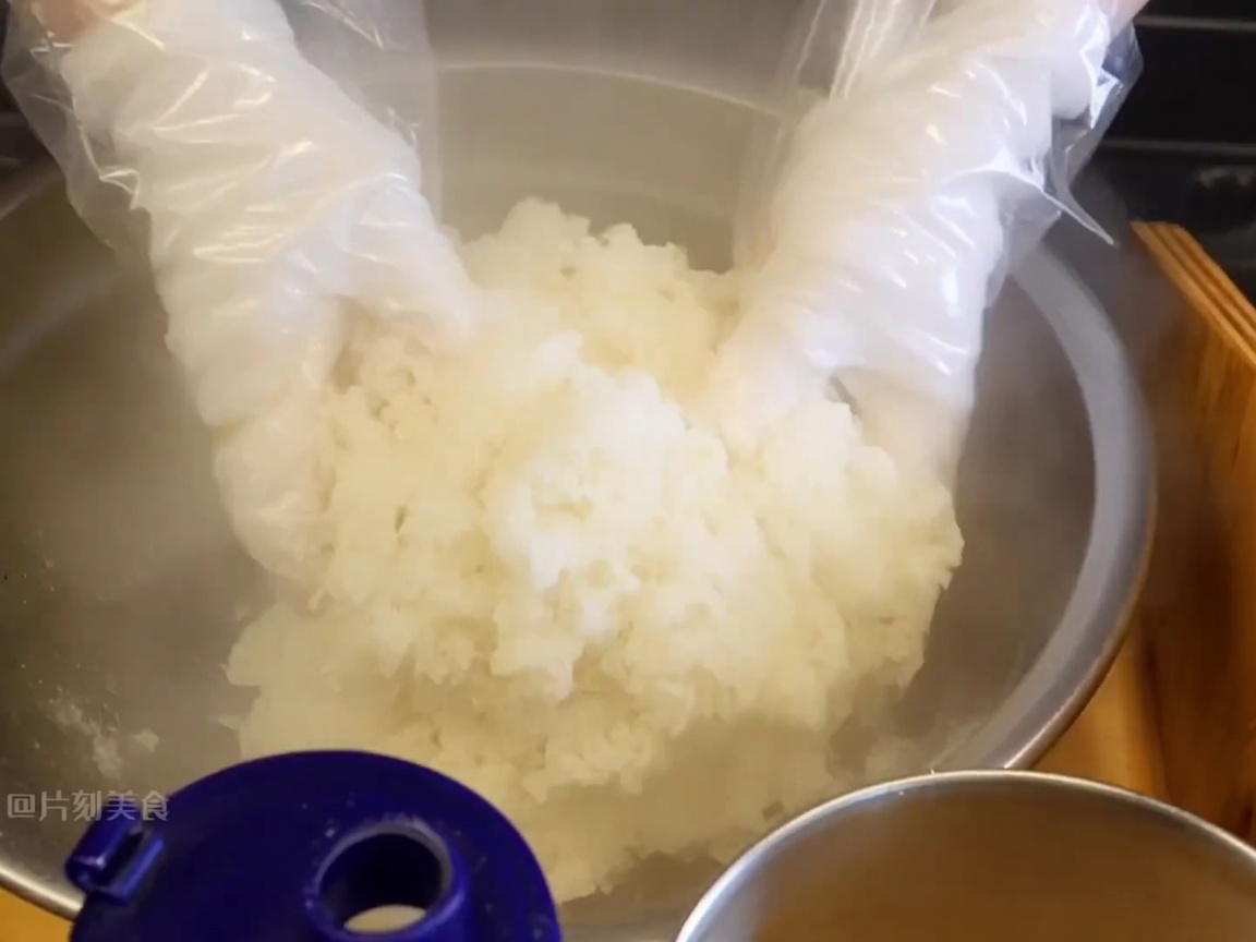 Manos con guantes mezclando arroz pegajoso humeante en un bol grande para incorporar los condimentos.