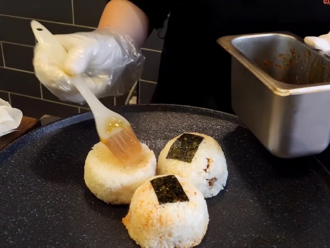 Bolas de arroz sobre una plancha caliente siendo pinceladas con un glaseado marrón rico por una mano enguantada.