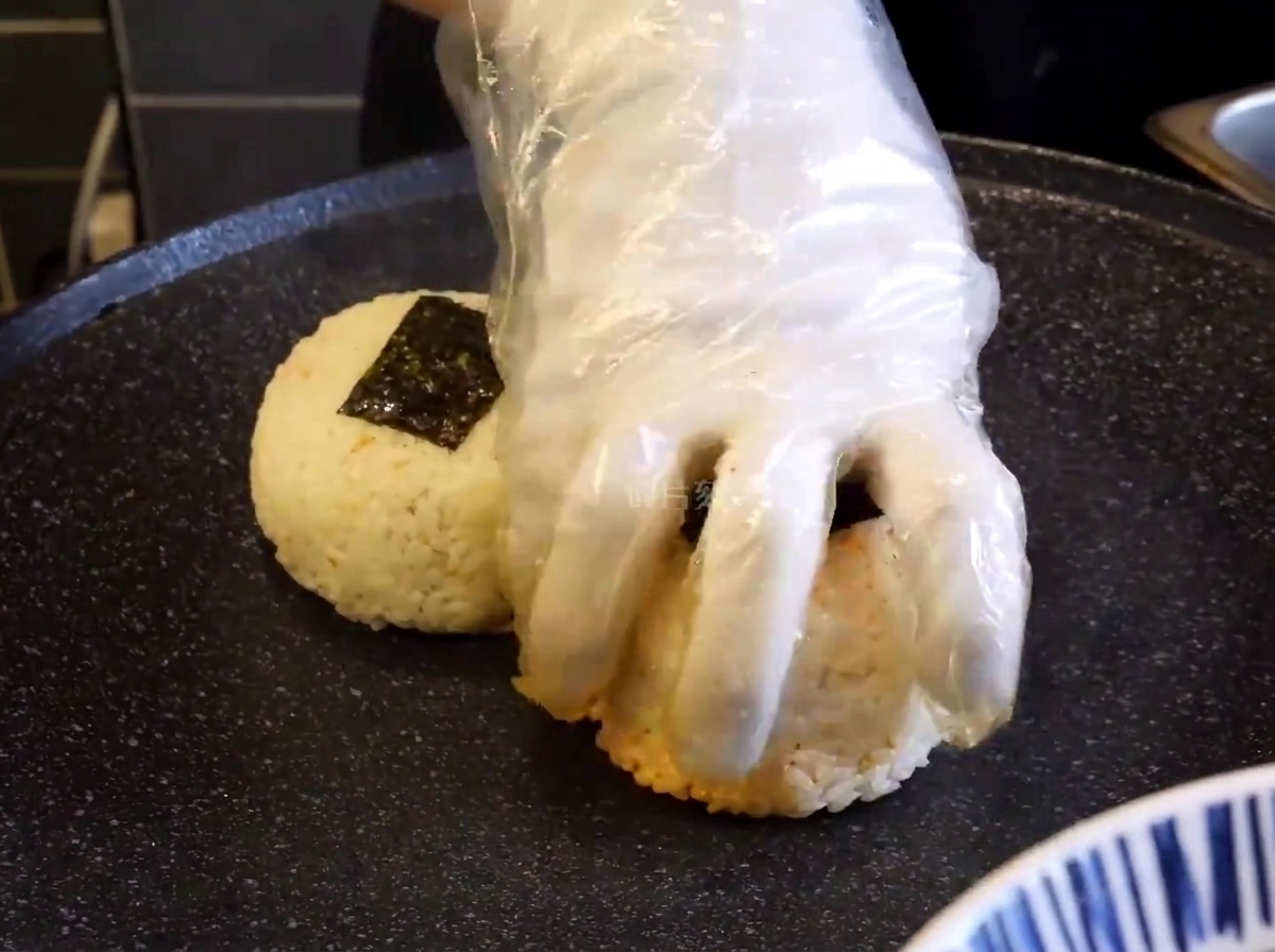 Primer plano de una mano enguantada presionando bolas de arroz sazonadas sobre una plancha negra caliente para sellarlas.
