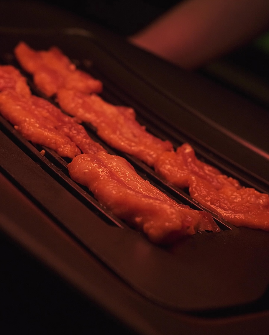 Rodajas de panceta marinada chisporroteando en una superficie de parrilla eléctrica caliente.