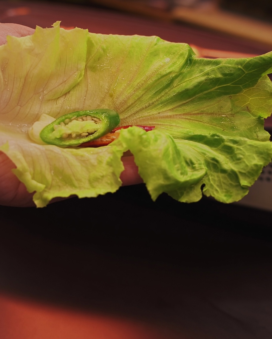 Una mano sosteniendo una hoja de lechuga fresca con un trozo de panceta a la parrilla y chile listo para el montaje.