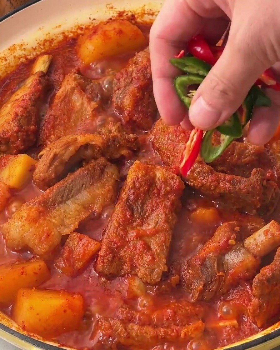 Una mano esparciendo rodajas de chile verde y rojo sobre la superficie del estofado terminado.