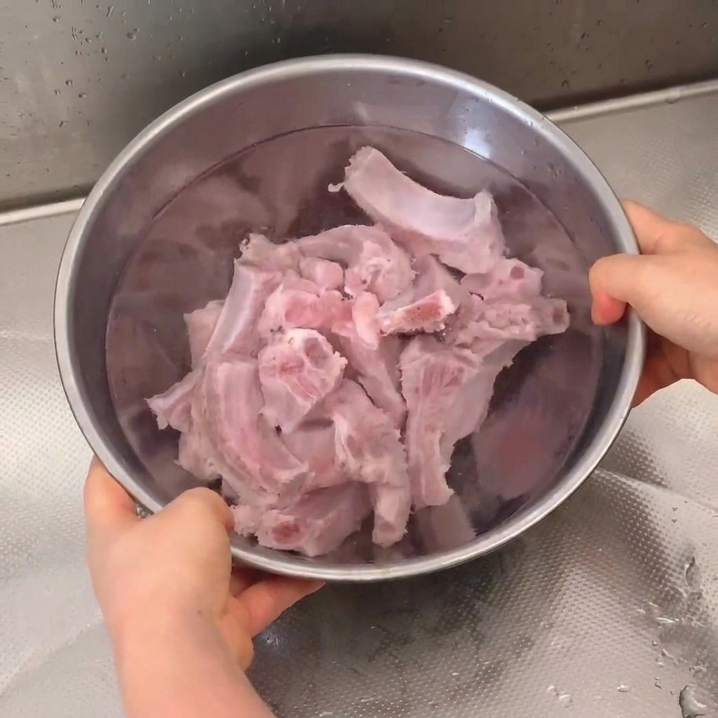 Costillas de cerdo frescas remojándose y siendo enjuagadas en un bol de acero inoxidable bajo el grifo.