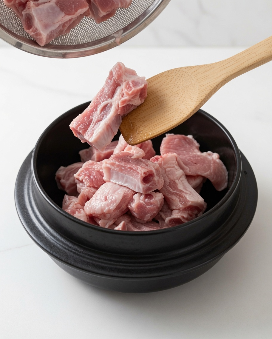 Costillas de cerdo sofriéndose en una olla con aceite y azúcar blanca usando una cuchara de madera.