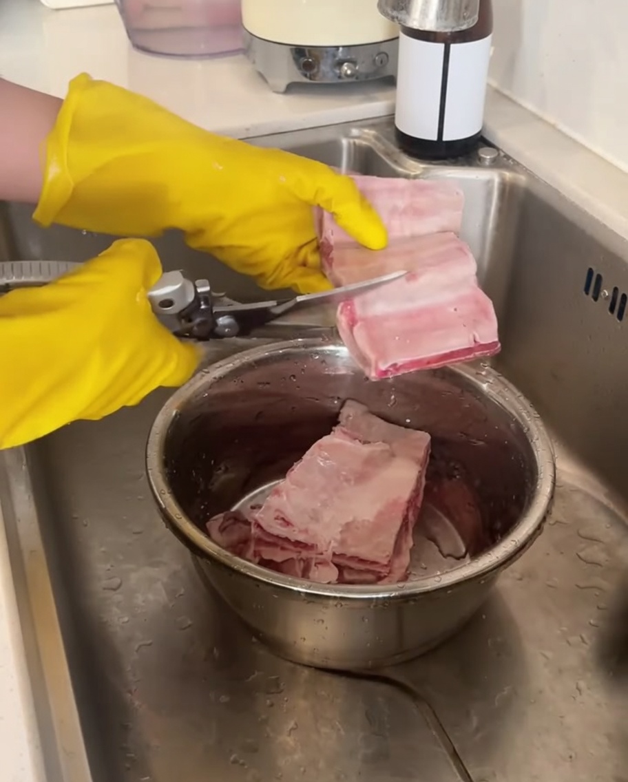 Un par de manos con guantes amarillos usando tijeras de cocina para cortar costillas de res crudas sobre un tazón de metal.