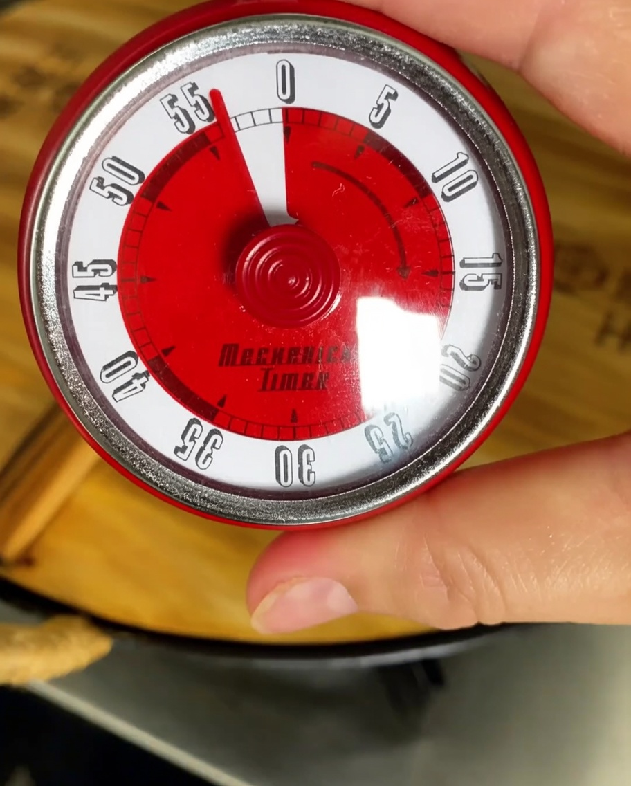 Un temporizador de cocina ajustado a una hora con una olla cocinando a fuego lento en el fondo.