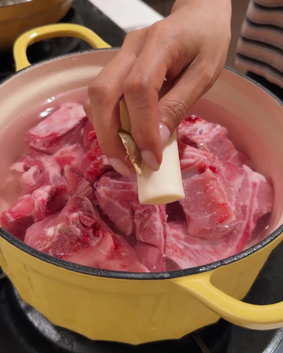 Una mano colocando un trozo de jengibre en una olla que contiene espinazo de cerdo y agua.