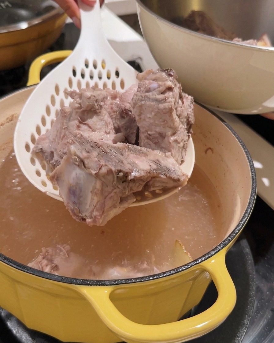 Un colador levantando trozos de espinazo de cerdo blanqueado fuera de una olla.