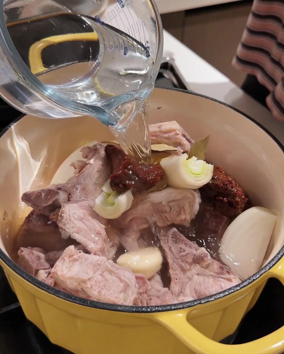 Agua caliente siendo vertida desde una taza medidora en la olla con cerdo y condimentos.