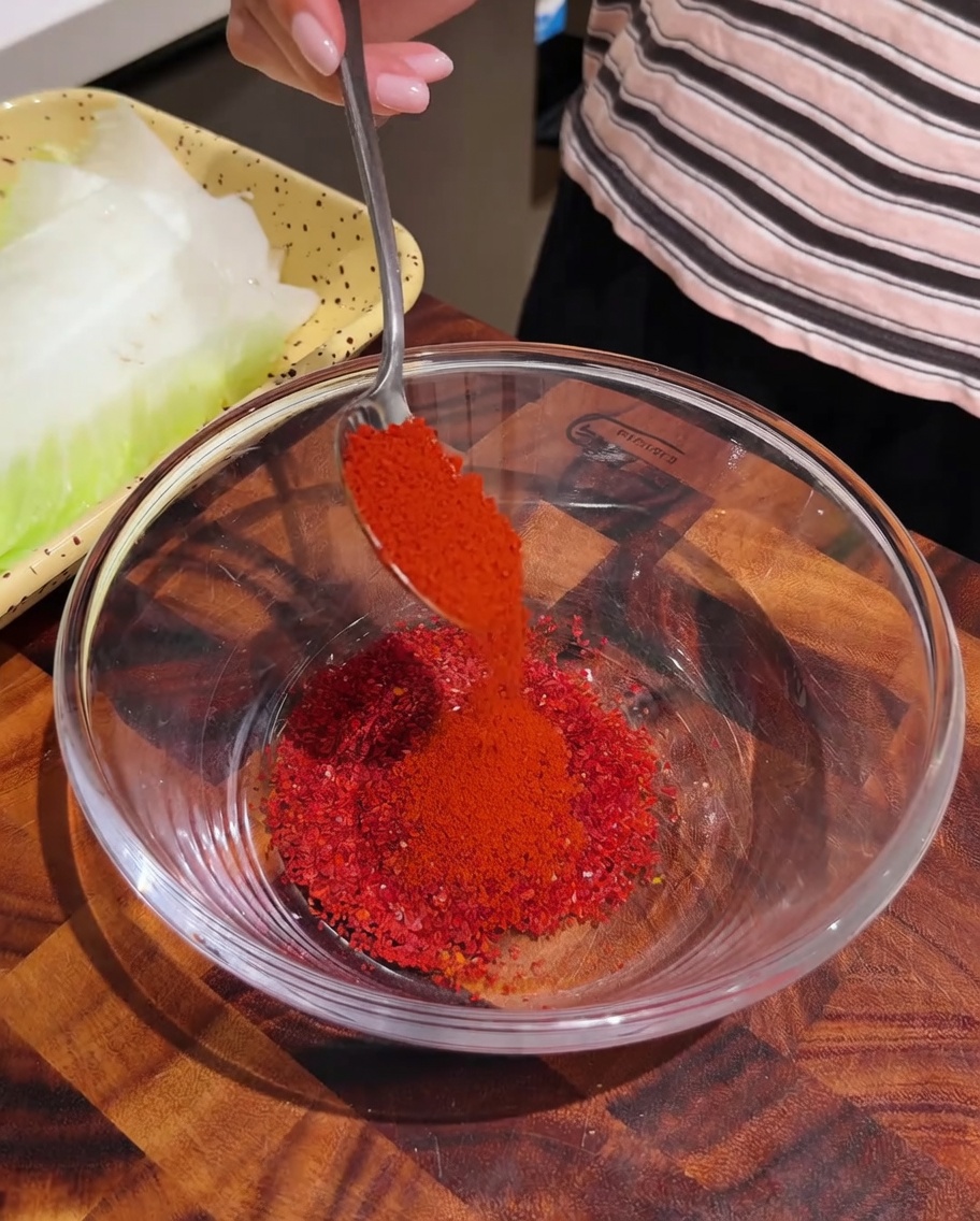 Polvo de chile rojo siendo servido con cuchara en un bol de cristal transparente que contiene otras especias de condimento.