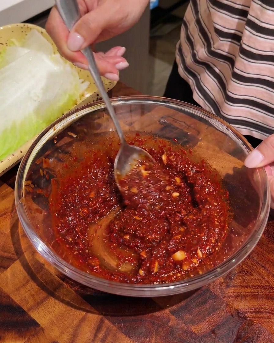Tiras desgarradas de col napa blanqueada siendo mezcladas con una salsa de sazonamiento roja en un bol de cristal.