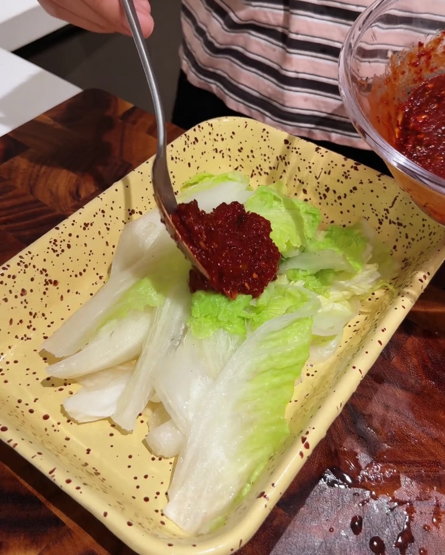 Cuchara aplicando pasta de sazonamiento roja sobre una bandeja de hojas de col fresca.
