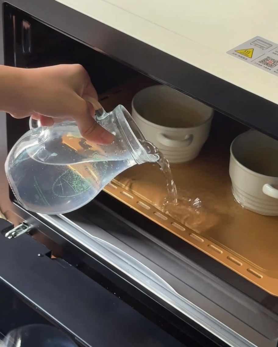 Agua tibia vertiéndose desde una jarra de cristal en una bandeja de horno que contiene ramequines de cerámica.