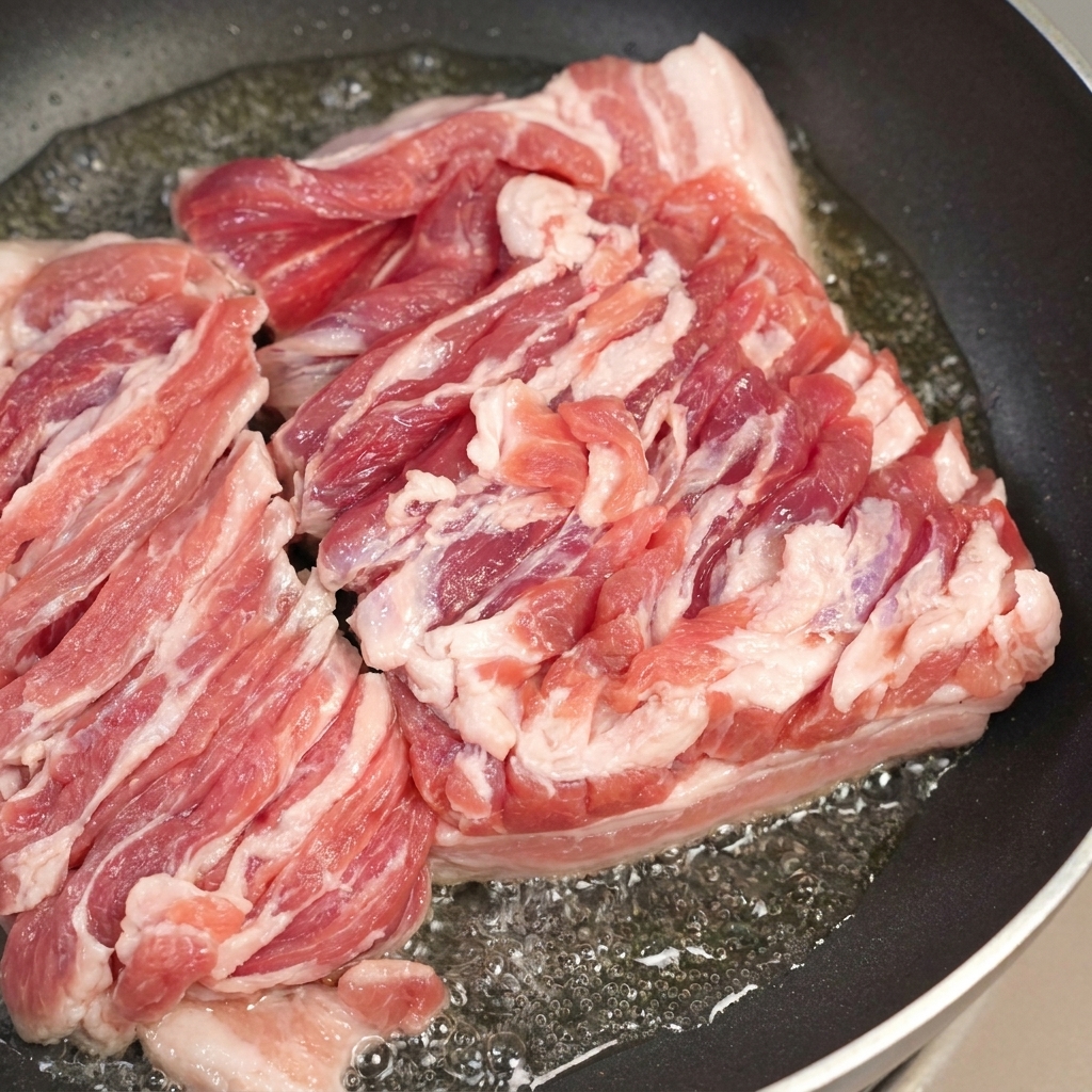 Una sartén con panceta de cerdo chisporroteando y una cuchara retirando el exceso de grasa derretida.