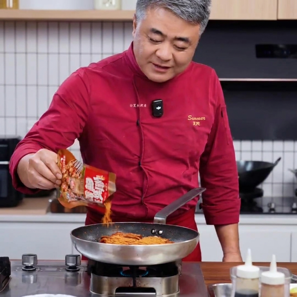 Chef con abrigo rojo vertiendo mezcla de especias para barbacoa desde una bolsa en una sartén con cerdo dorado.
