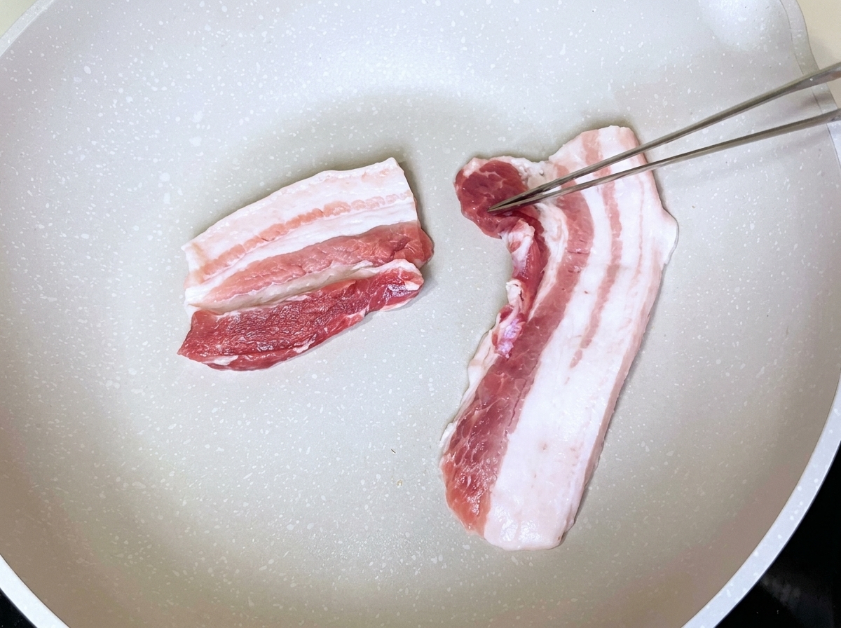 Rebanadas de panceta de cerdo colocadas en una sartén blanca seca siendo manipuladas con pinzas metálicas.