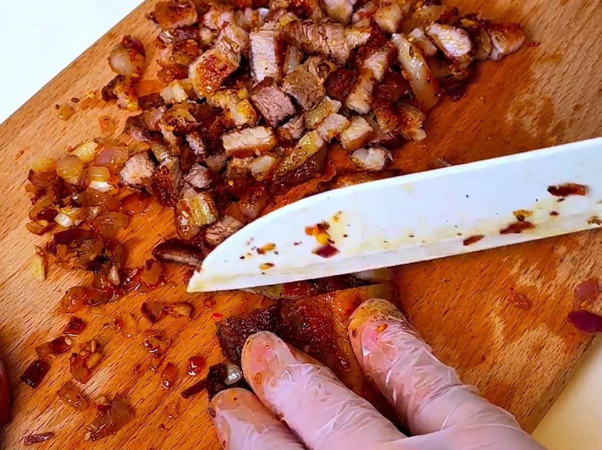 Un cuchillo picando cerdo barbacoa cocido y cebollas en una tabla de madera, con manos usando guantes protectores.