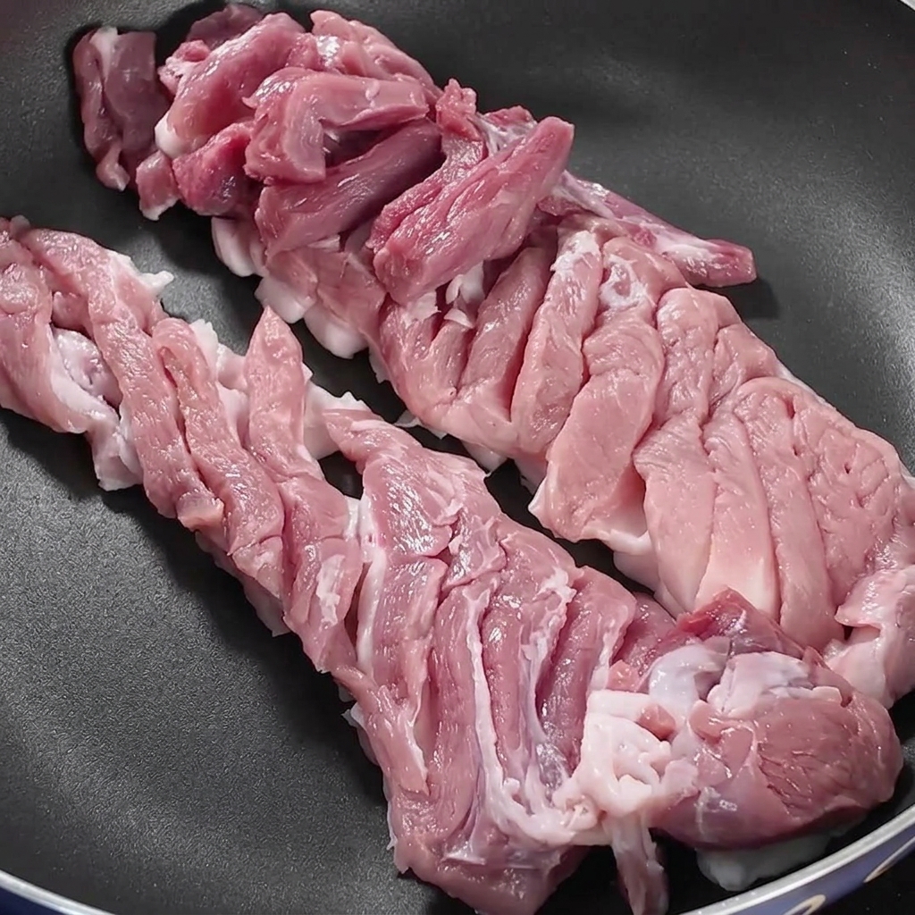 Rebanadas de panza de cerdo dispuestas ordenadamente dentro de una sartén oscura sobre un quemador de estufa.