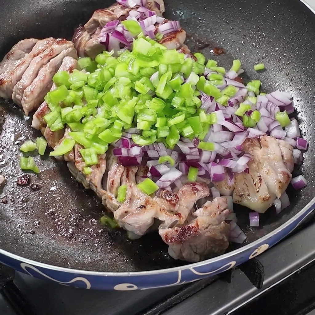 Una sartén llena de panza de cerdo frita cubierta con montones de cebollas picadas, pimientos verdes y especias secas.