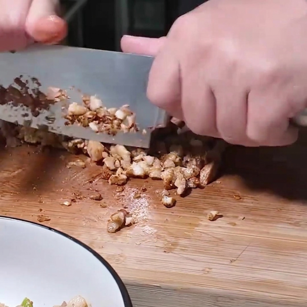 Una persona usando un cuchillo grande para picar finamente trozos de cerdo cocido en una tabla de madera junto a un bol para mezclar.