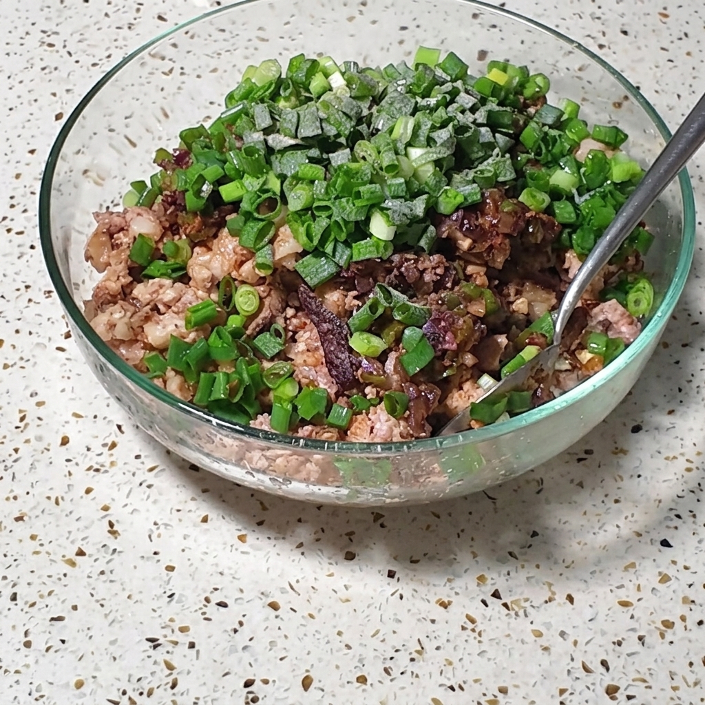 Un bol de vidrio que contiene una mezcla de cerdo finamente picado y huevos, cubierto con cebollas verdes frescas y condimentos, siendo revuelto con una cuchara de metal.