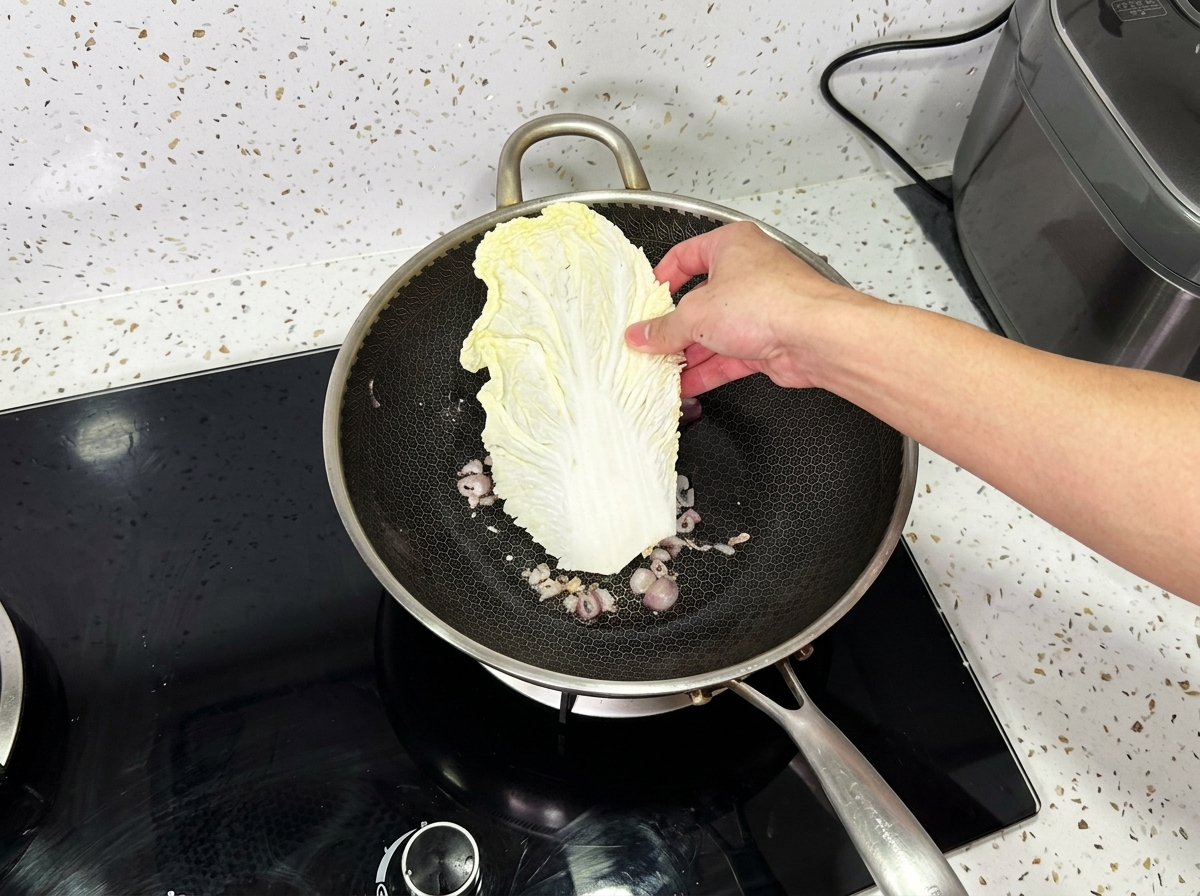 Una mano colocando una hoja grande de col napa en un wok que contiene cebollas cocinadas.