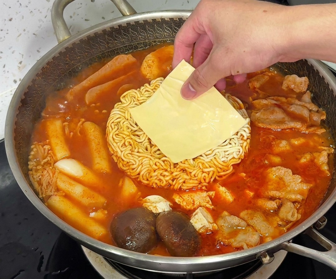 Una rebanada de queso amarillo derritiéndose sobre los fideos ramen en una olla humeante de Budae Jjigae.