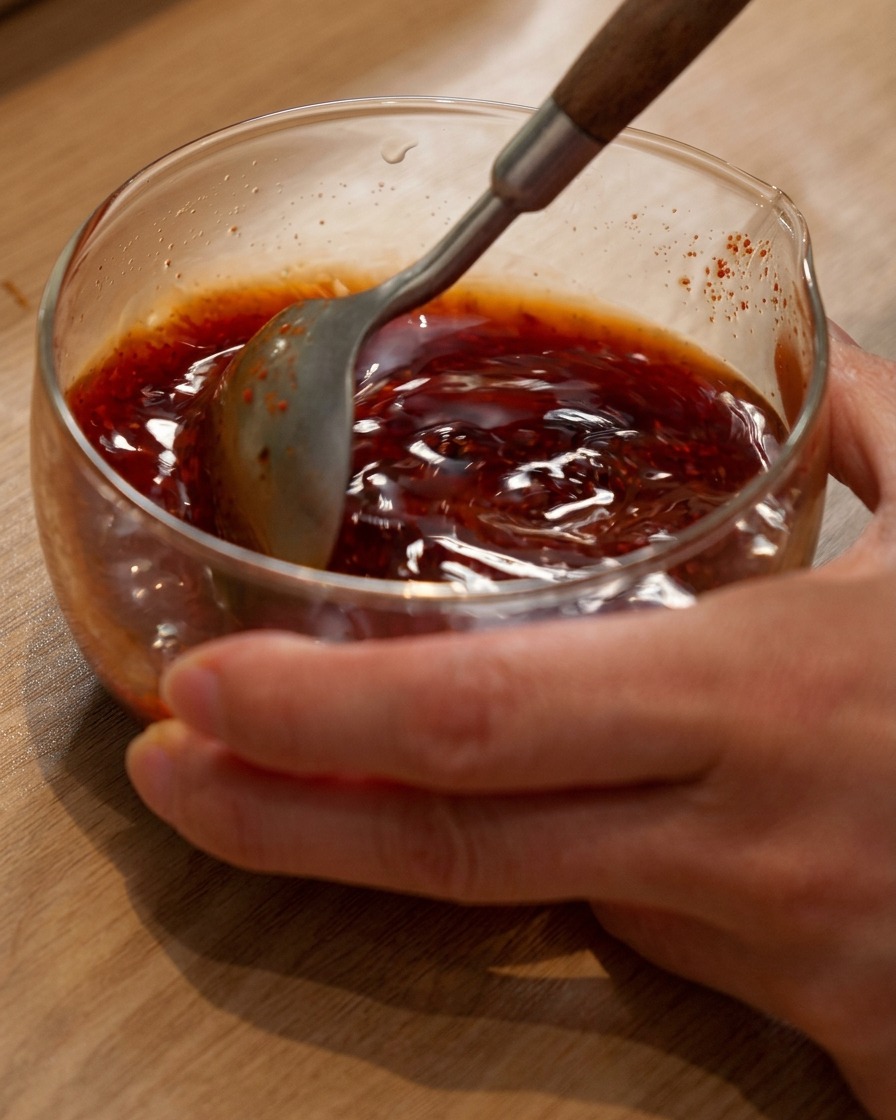 Una cuchara removiendo una mezcla de salsa para hot pot de color marrón rojizo en un bol de cristal.