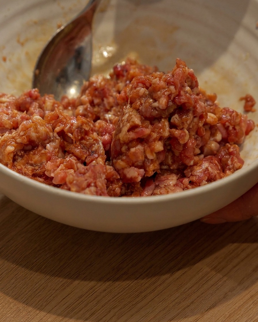 Carne de cerdo picada siendo marinada en un bol blanco con una cuchara.