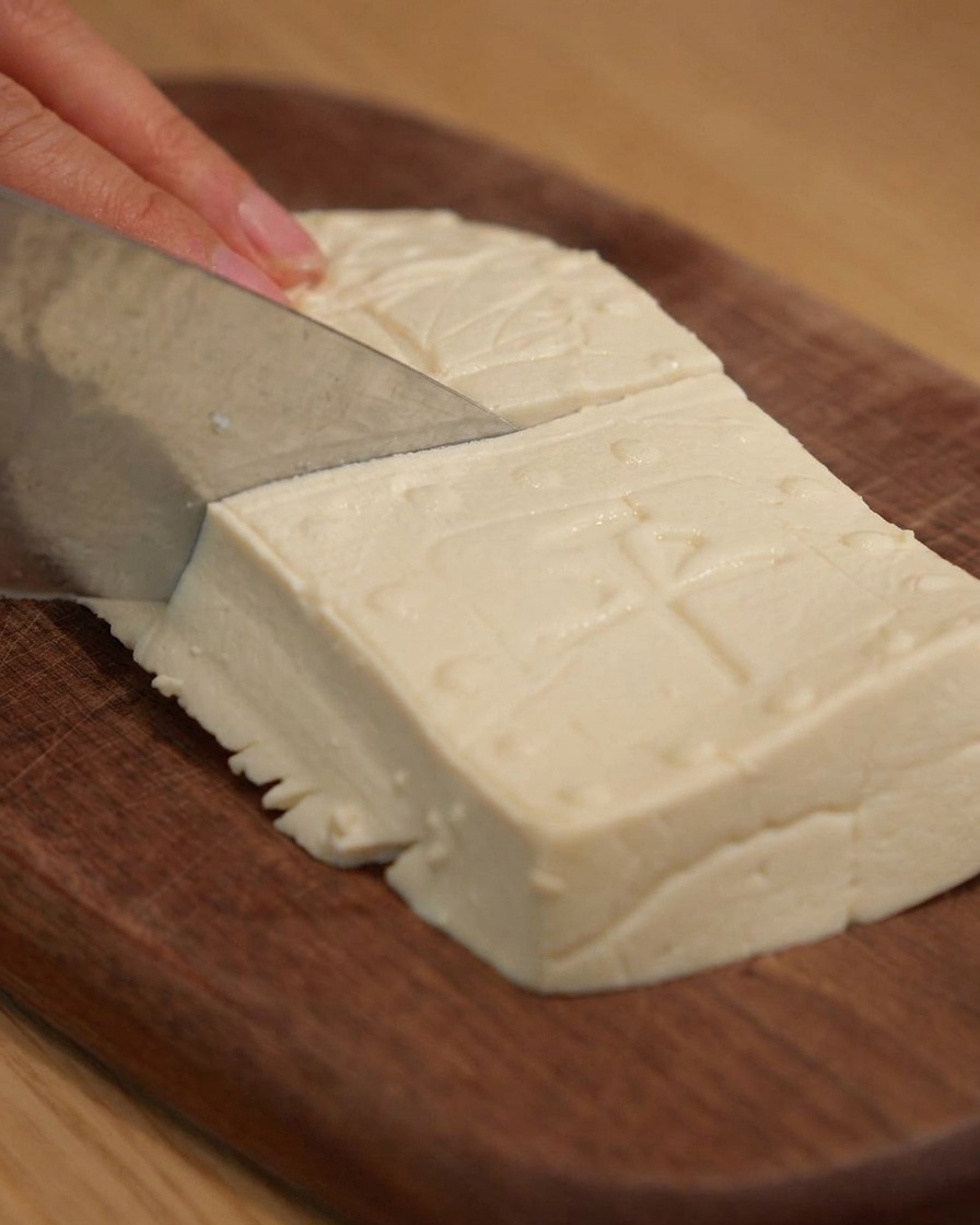Un cuchillo afilado cortando tofu firme en bloques rectangulares limpios sobre una tabla de cortar de madera.