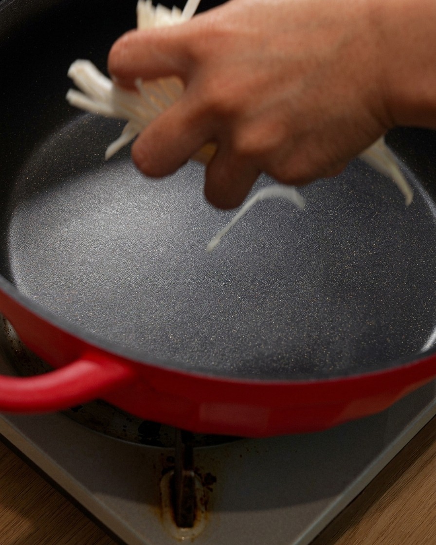 Cebollas cortadas en tiras siendo añadidas en el fondo de una olla roja.