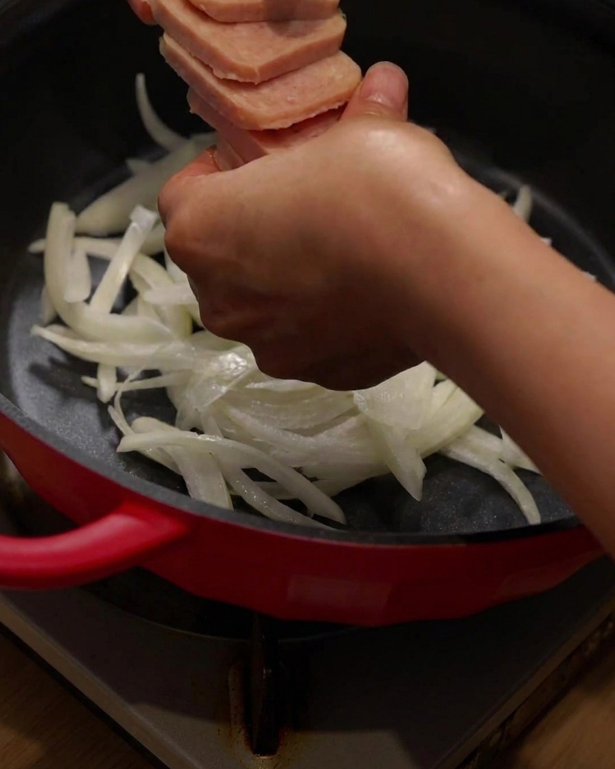 Organizando carne en conserva cortada y tocino encima de las cebollas en la olla de cocción roja.