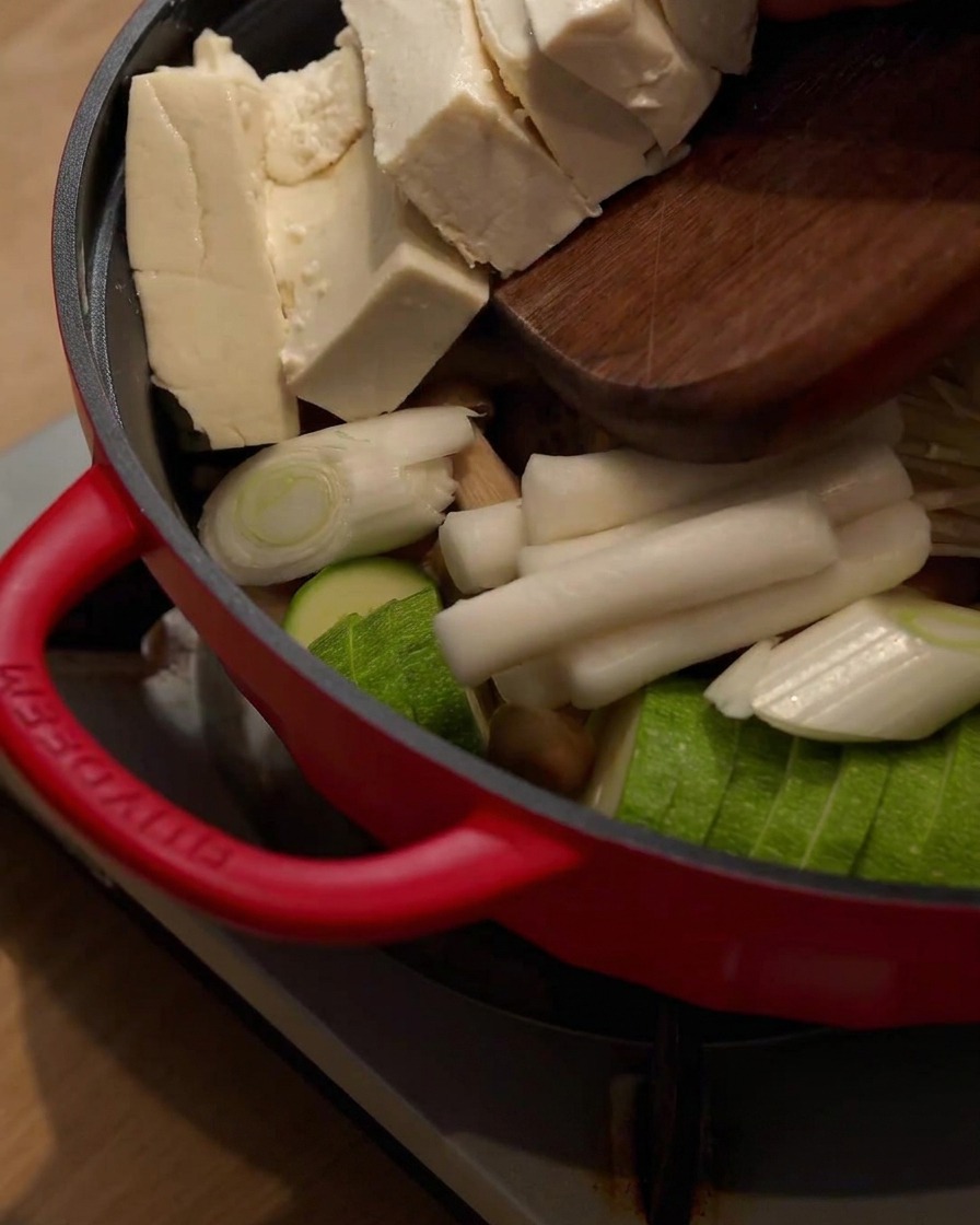 Bloques de tofu siendo deslizados desde una tabla de cortar de madera hacia el hot pot que contiene otros ingredientes.