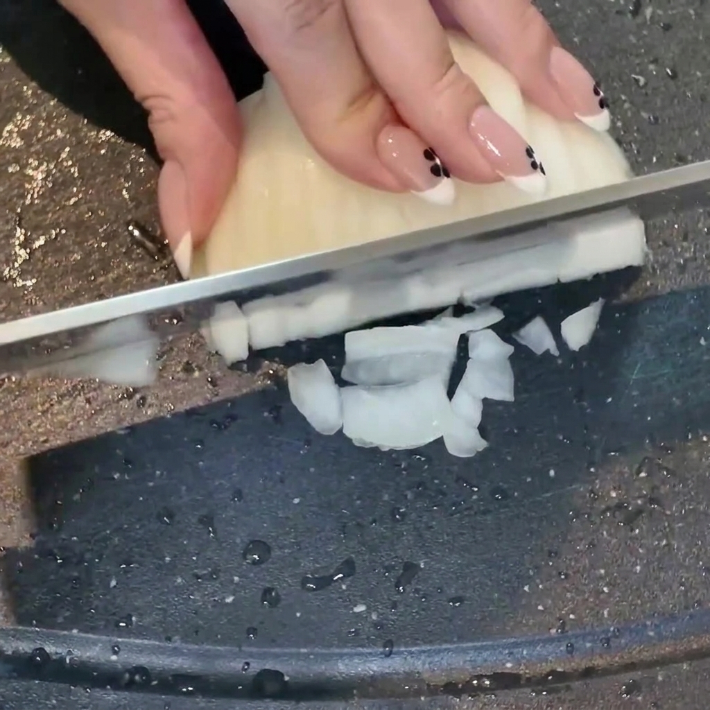 Una mano usando un cuchillo para cortar una cebolla blanca en dados sobre una tabla de cortar oscura, con ajo picado cerca.