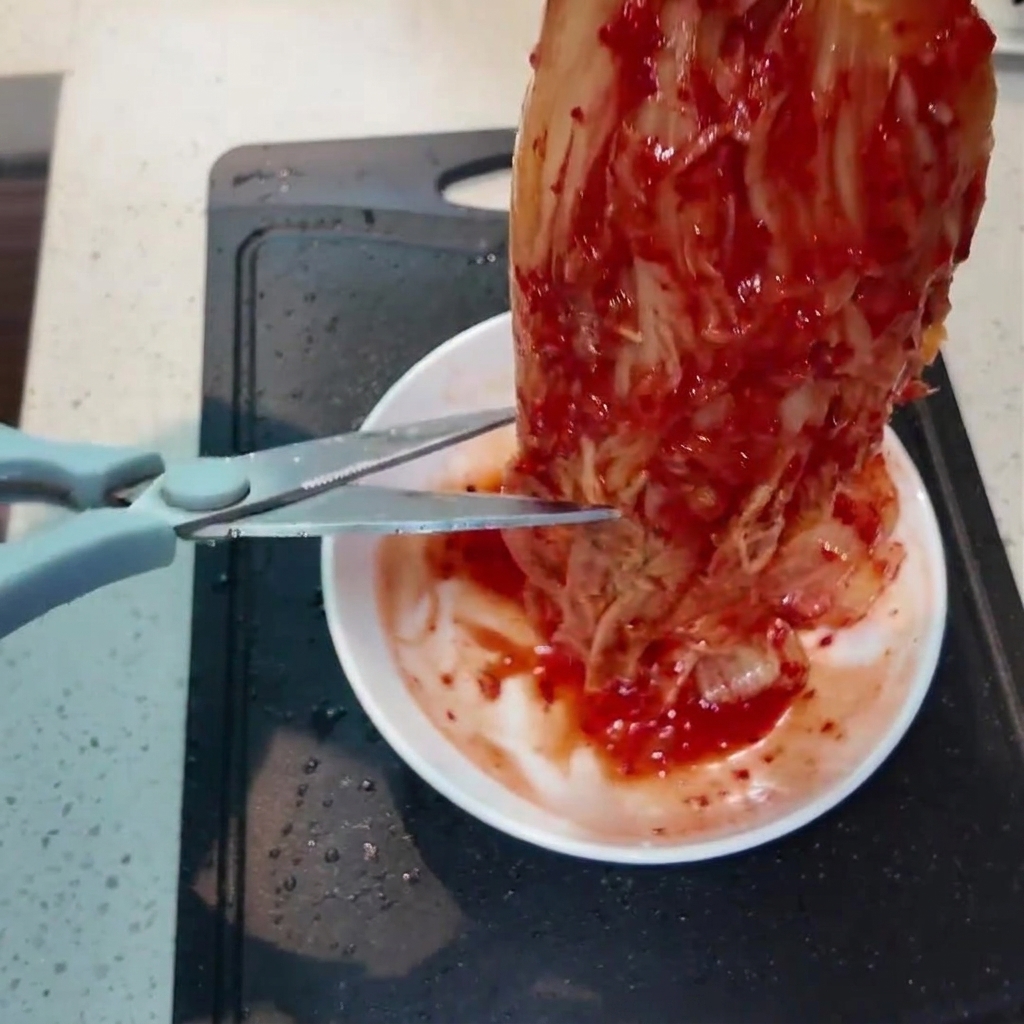 Un trozo de kimchi rojo vibrante siendo cortado en trozos más pequeños con tijeras de cocina sobre un plato blanco pequeño.