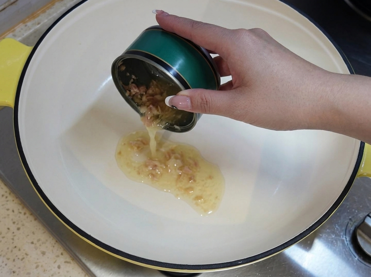 Una mano sosteniendo una lata de atún verde vacía, vertiendo su aceite residual en una olla blanca caliente en la estufa.