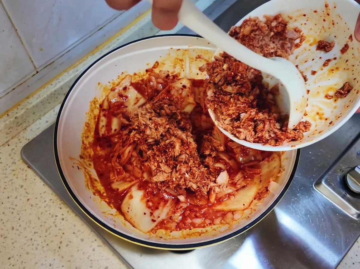 Mezcla de atún en lata siendo retirada de un tazón blanco a la sartén con kimchi usando una cuchara blanca.