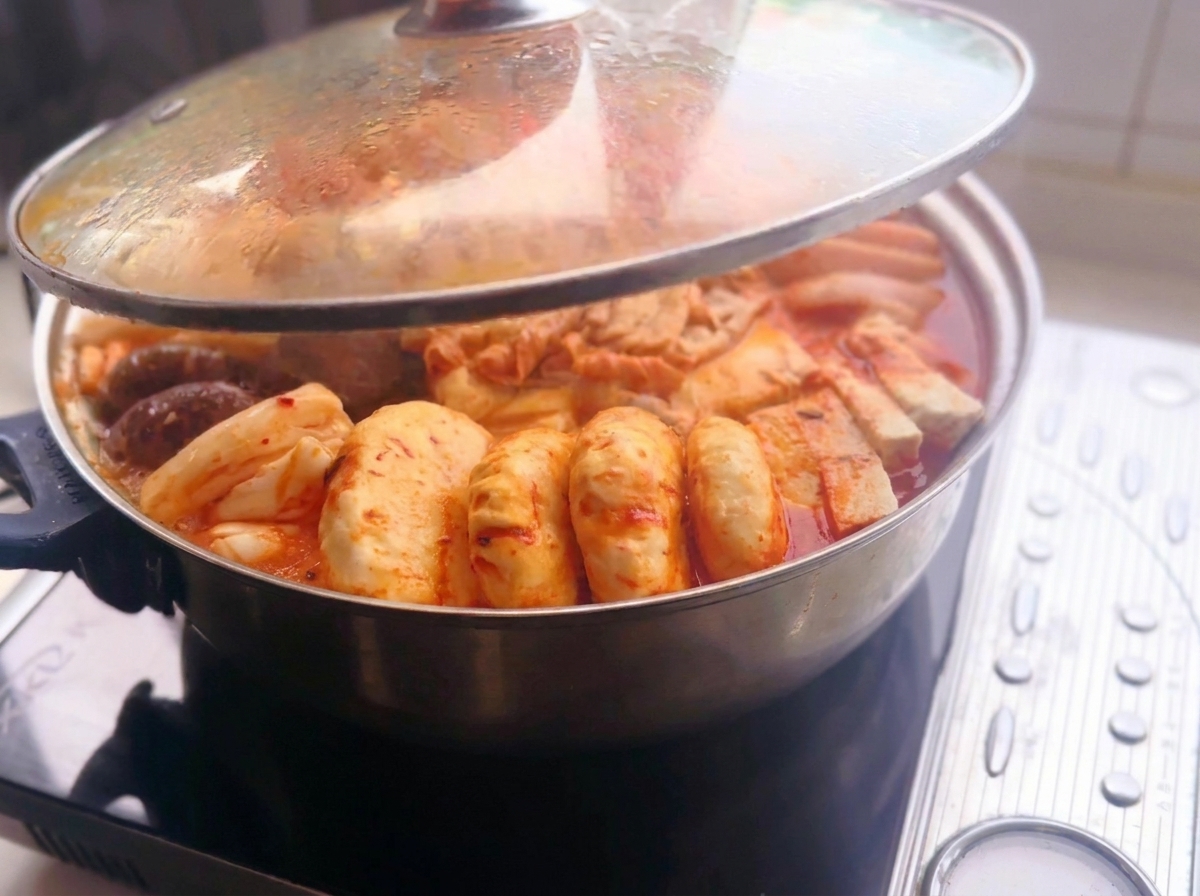 Una tapa de vidrio siendo levantada de una olla caliente de metal llena de ingredientes dispuestos como carne en rodajas, tofu, champiñones, pasteles de arroz y albóndigas en un caldo rojo picante en una cocina de inducción.