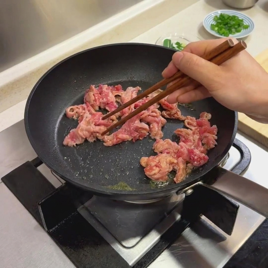 Rodajas de ternera marinada siendo cocinadas en una sartén negra usando palillos de madera.