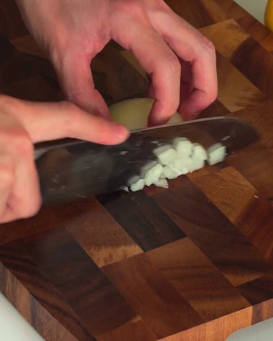 Manos sosteniendo un cuchillo y picando finamente una cebolla blanca en una tabla de madera.
