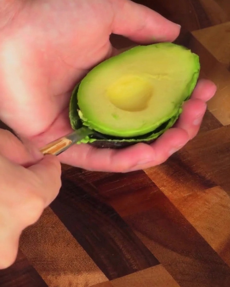 Una persona usando una cuchara para sacar la pulpa verde de la mitad de un aguacate.