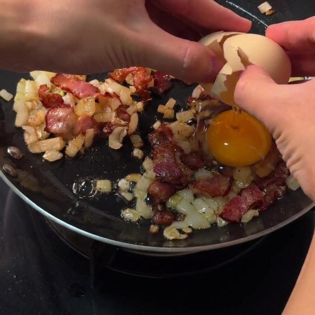 Manos rompiendo un huevo crudo directamente en un espacio circular despejado entre tocino y cebollas en una sartén.