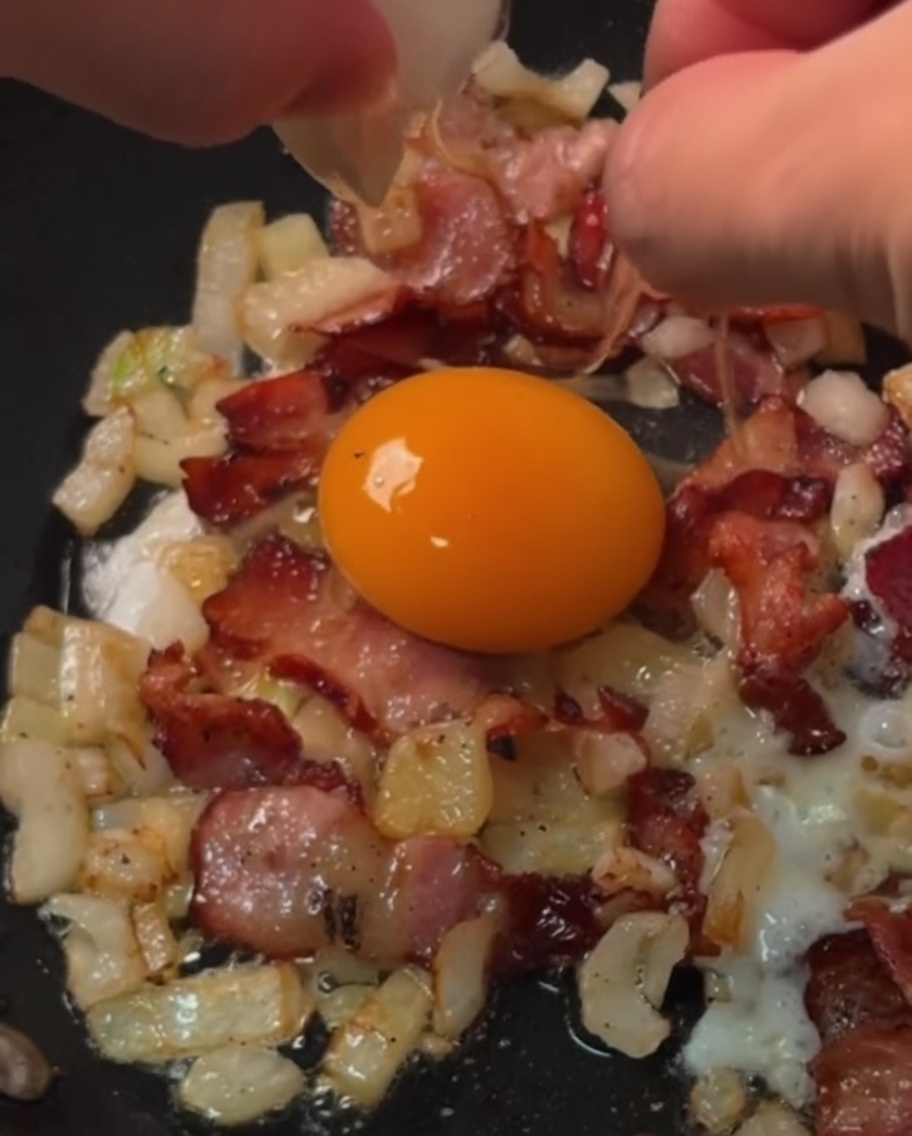 Huevos frescos siendo rotos en anillos individuales de tocino y cebolla cocinándose en una sartén.