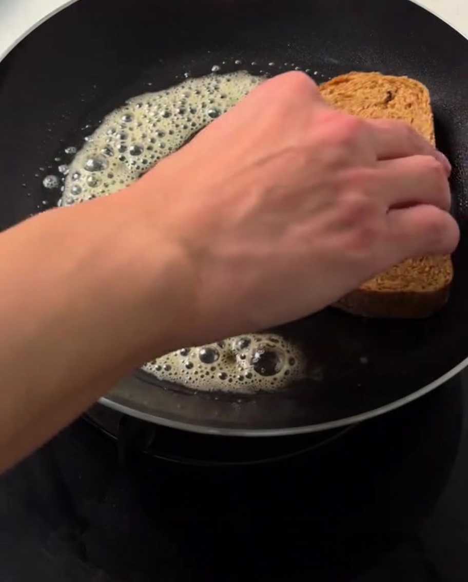 Una mano colocando una rebanada de pan integral sobre mantequilla derretida y burbujeante en una sartén caliente.