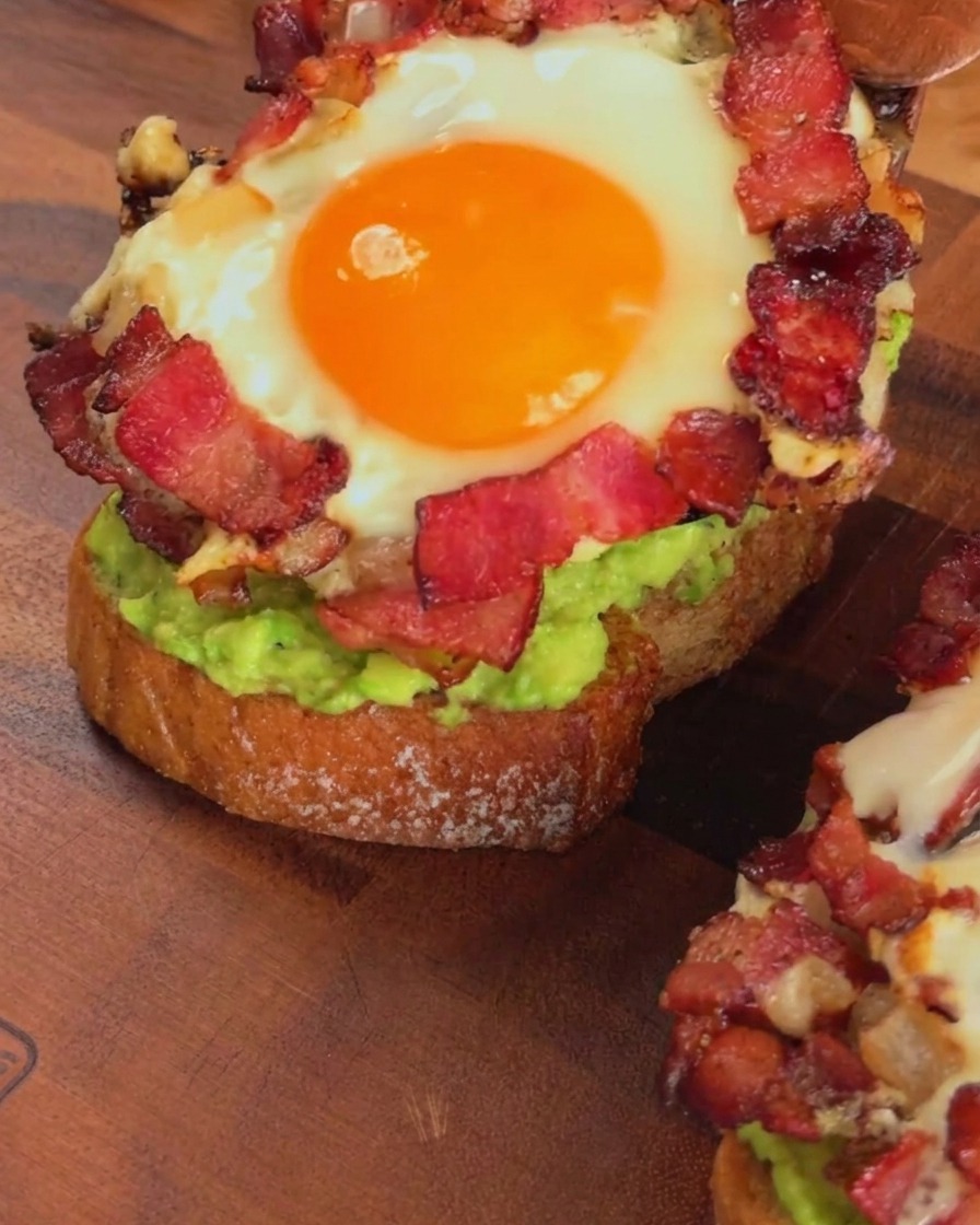 Un anillo de tocino y huevo cocido siendo colocado sobre una tostada de aguacate con una espátula de madera.