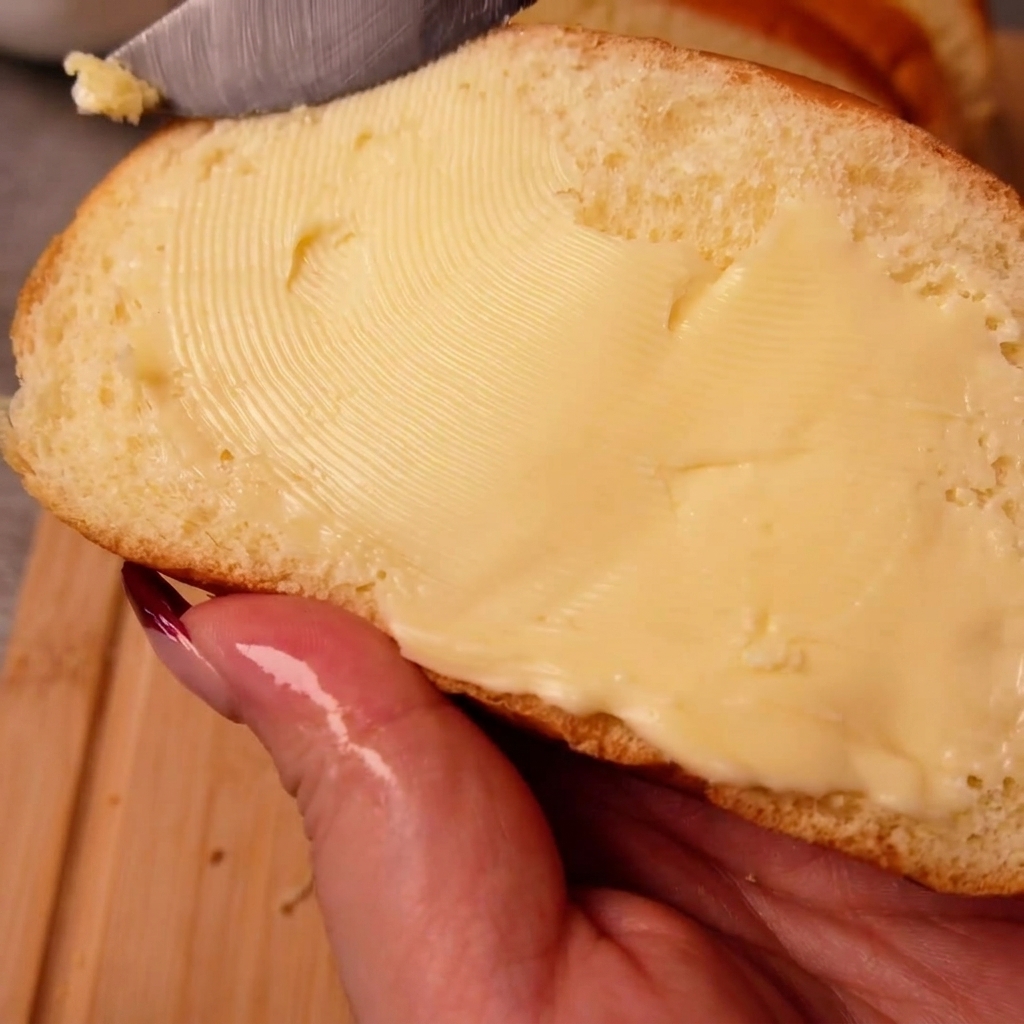 Una mano sosteniendo una rebanada de pan blanco mientras esparce mantequilla uniformemente sobre su superficie con un cuchillo.